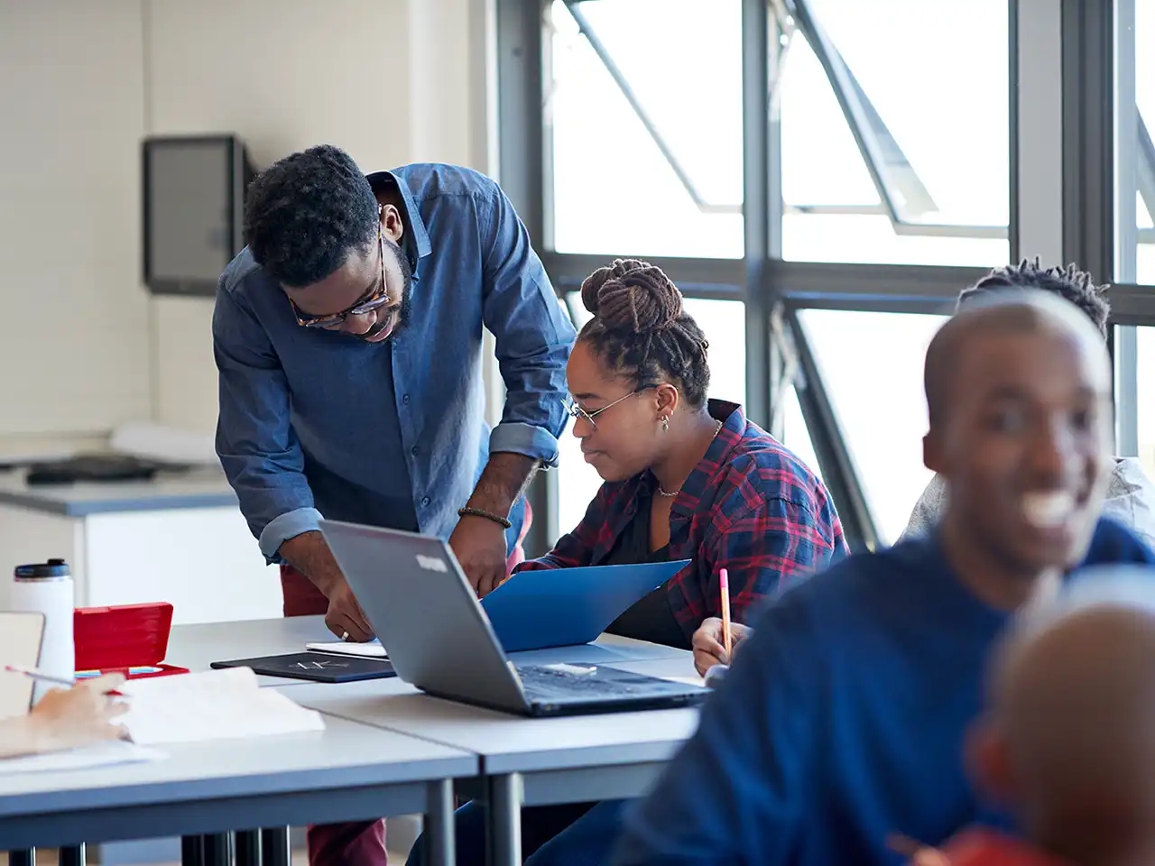 Enseignant se penchant sur le bureau pour aider un élève à utiliser son ordinateur portable dans une salle de classe.