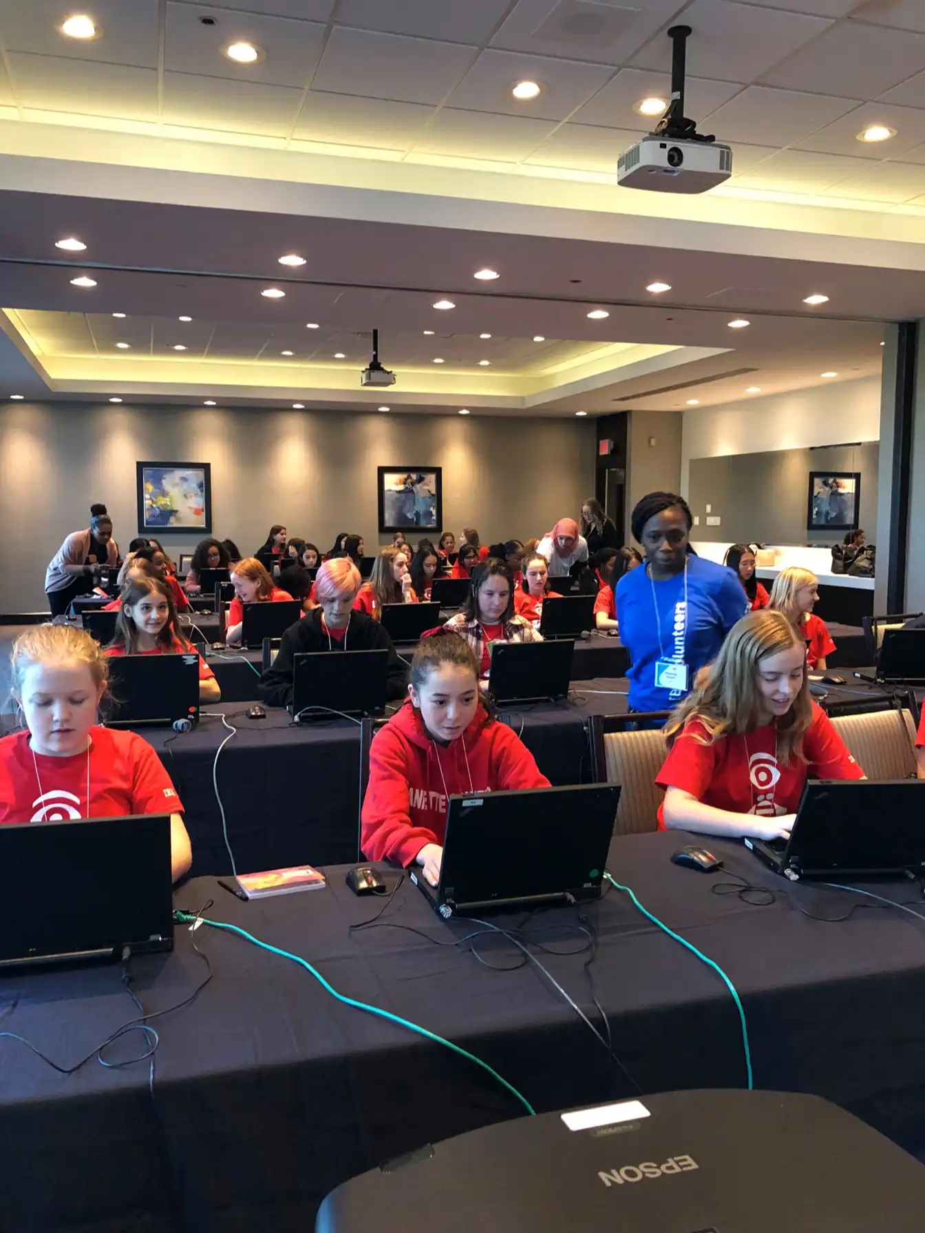 Klassenzimmer mit CyberDay4Girls-Teilnehmerinnen, die an Laptops arbeiten, und Mentoren, die den Schülerinnen helfen