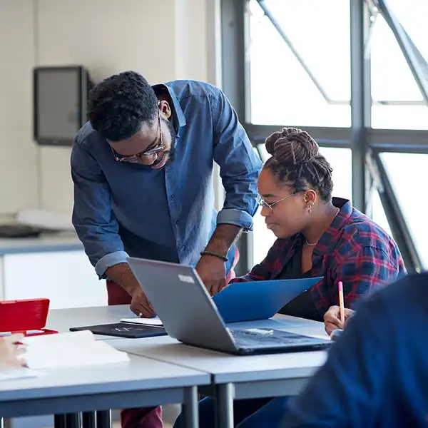 Teacher leaning over desk to assist student at laptop in classroom